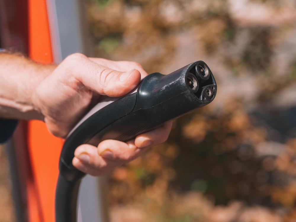 Main tenant une prise de recharge de véhicule électrique devant une borne